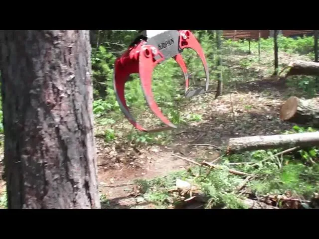 Close-up of a red MARK II grapple attachment open in a pine woodlot beside cut logs