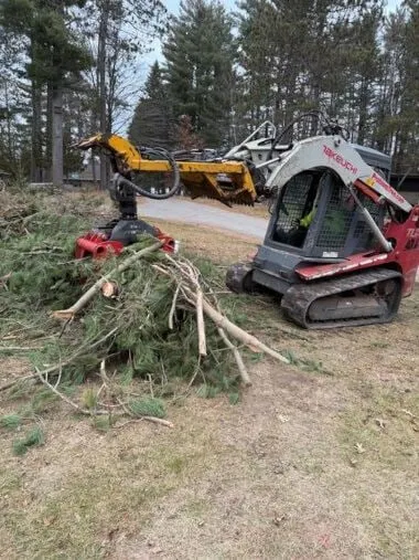 Tracked skid steer with a mulching attachment beside a pile of cleared brush and branches
